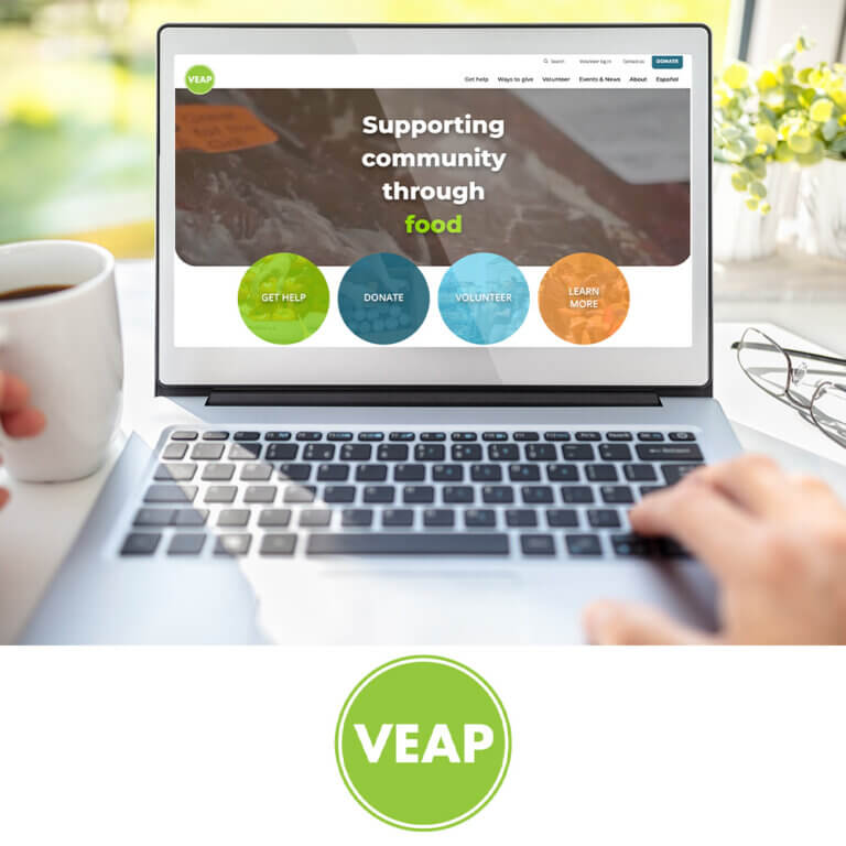 A person using a laptop to view the VEAP website. The screen displays VEAP’s homepage with the message “Supporting community through food” and four colorful circular buttons labeled “Get Help,” “Donate,” “Volunteer,” and “Learn More.” A coffee cup, glasses, and bright natural light complete the scene. Below the laptop image is the green VEAP logo with white bold lettering inside a circle.