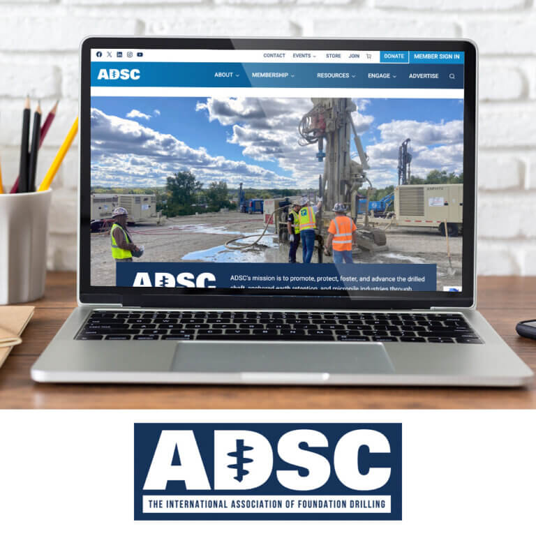 A laptop on a wooden desk displays the redesigned ADSC website featuring workers operating drilling equipment on a construction site. Below the laptop, the ADSC logo reads ‘The International Association of Foundation Drilling.’