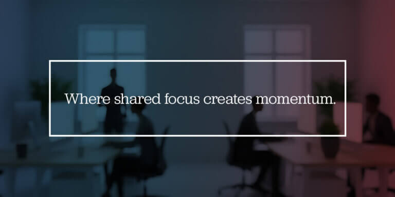 Blurry office scene with silhouetted people working at desks; centered white rectangular frame contains the phrase ‘Where shared focus creates momentum.’